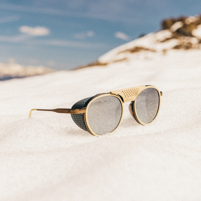 Round sunglasses with gold-tone metal frames, perforated side details, and dark lenses resting in white snow with a blurred blue sky and mountain background.
