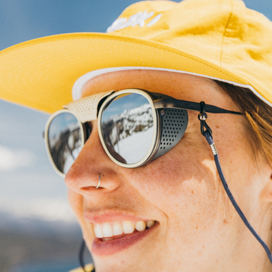 Close-up of a person outdoors wearing round sunglasses with a black chin strap and a yellow sun hat. The sunglasses have a light beige front frame with dark gray perforated side panels and mirrored lenses.