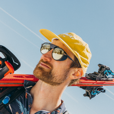 A man outdoors wearing a yellow cap, black sunglasses, and a patterned shirt, with red skis resting over his shoulders against a blue sky.