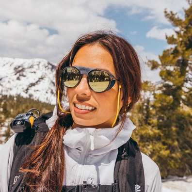 A woman outdoors in a snowy mountain setting wearing dark sunglasses with gold-tinted lenses, a white hooded jacket, and a black backpack.