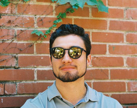 Man wearing sunglasses in front of a brick wall with greenery