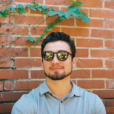 Man wearing sunglasses in front of a brick wall with greenery