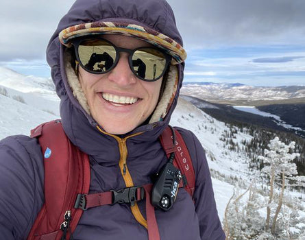 Person in winter clothing with a backpack and sunglasses, standing on a snowy mountain.