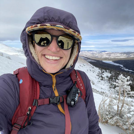 Person in winter clothing with a backpack and sunglasses, standing on a snowy mountain.