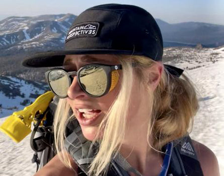 Person on a snowy mountain with goggles and a cap, with mountains in the background
