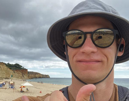 Person on a beach giving a thumbs-up with sunglasses and a wide-brimmed hat.