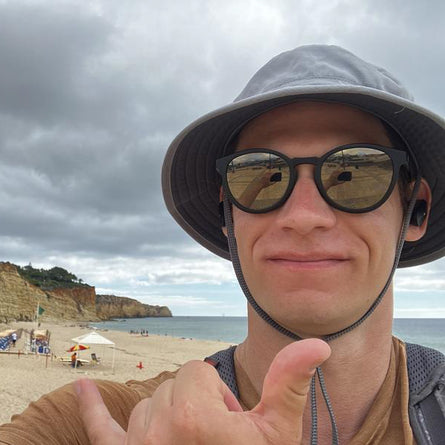 Person on a beach giving a thumbs-up with sunglasses and a wide-brimmed hat.