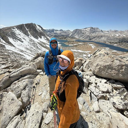 Two hikers on a rocky mountain summit with a scenic view of mountains and a lake.