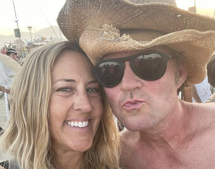 Two people posing for a selfie, one wearing a cowboy hat and sunglasses, with a blurred background.