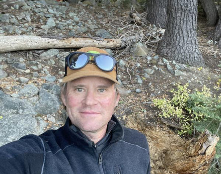 Person wearing a cap with sunglasses on top, standing in a forested area with trees and rocks.