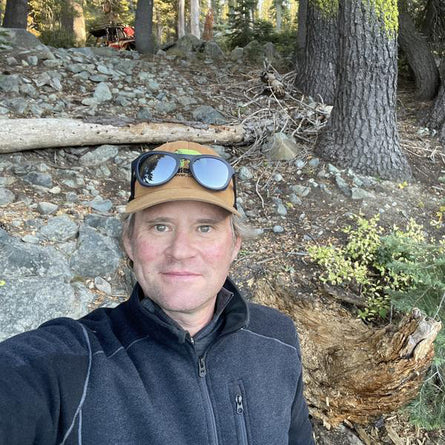Person wearing a cap with sunglasses on top, standing in a forested area with trees and rocks.