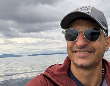 Man wearing sunglasses and a cap with a scenic background of water and mountains.