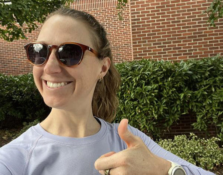 Person wearing sunglasses and a light purple shirt, giving a thumbs-up in front of a brick wall and greenery.