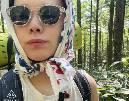Person wearing sunglasses and a colorful headscarf with a forest background