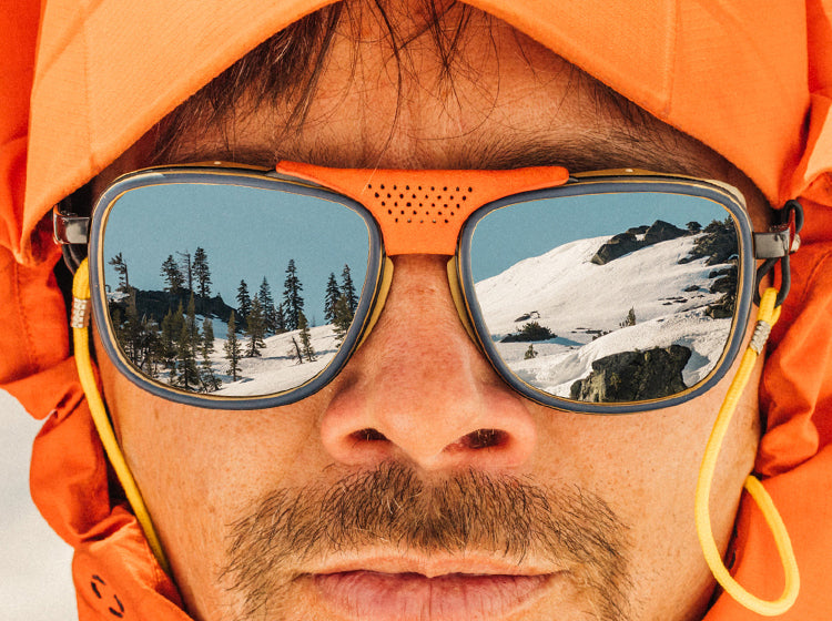 Man wearing sunski sunglasses with a snowy landscape reflection, wearing an orange jacket.