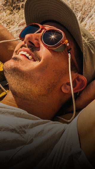 Man wearing a wide-brimmed hat and goggles, lying on the ground with a natural background.