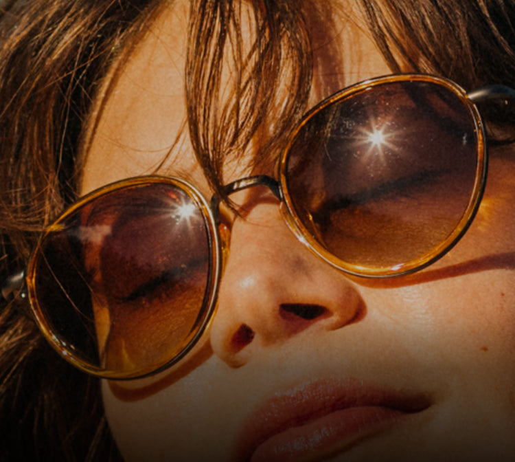 Close-up of a person wearing round sunglasses with a reflective sun in the background