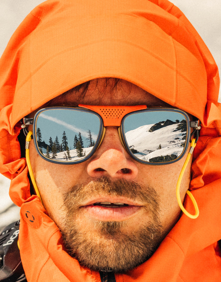 Person wearing an orange jacket and sunglasses with mountain reflection, snowy landscape