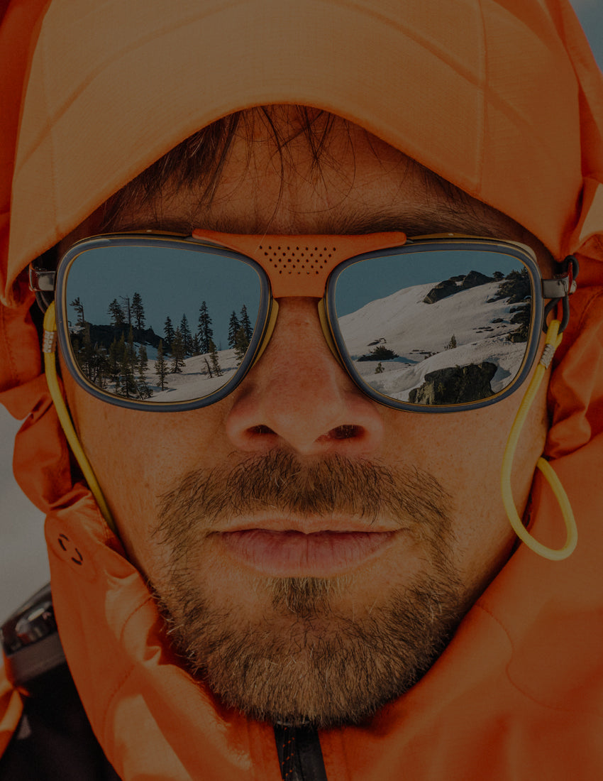 Person wearing orange goggles and jacket with a snowy mountain reflection.
