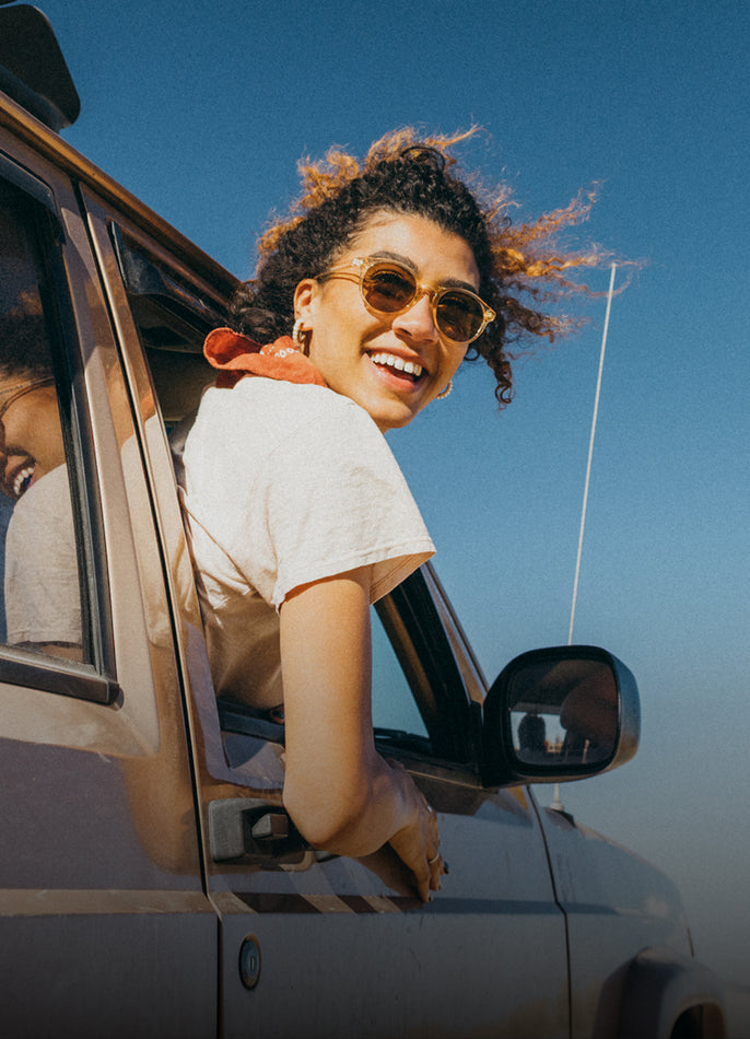 Woman with sunglasses smiling while leaning out of a car window against a blue sky.