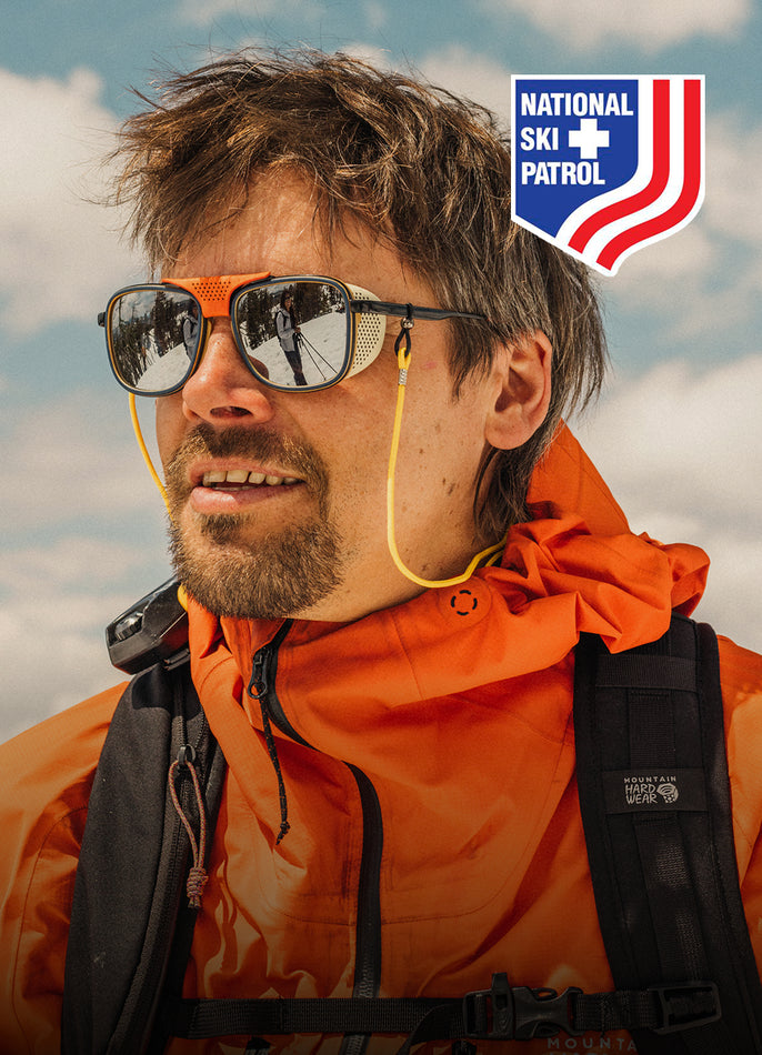 Person wearing an orange jacket and sunglasses with a National Ski Patrol logo in the corner.