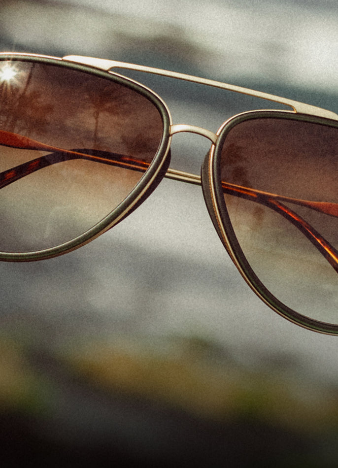 Aviator sunglasses with brown lenses and gold frame against a blurred natural background