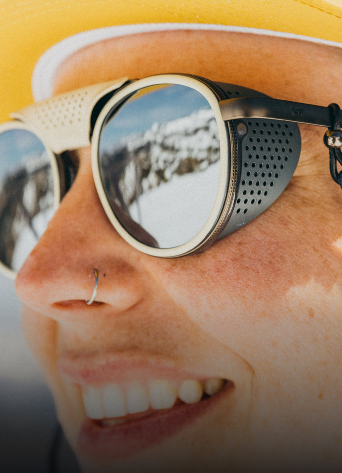 Person wearing reflective sunglasses with a snowy mountain reflection, wearing a yellow cap.