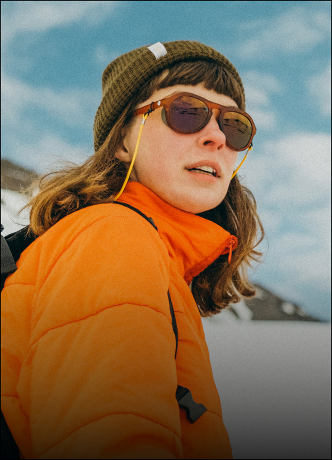 Person wearing an orange jacket and green beanie with sunglasses, standing outdoors against a blue sky.