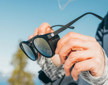 Person holding a pair of black sunglasses with a blurred outdoor background
