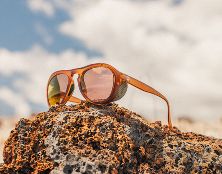 Sunglasses with orange frame and pink-tinted lenses on a rocky surface with a blurred natural background