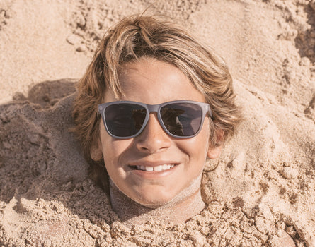 boy buried in the sand wearing sunski mini headland sunglasses