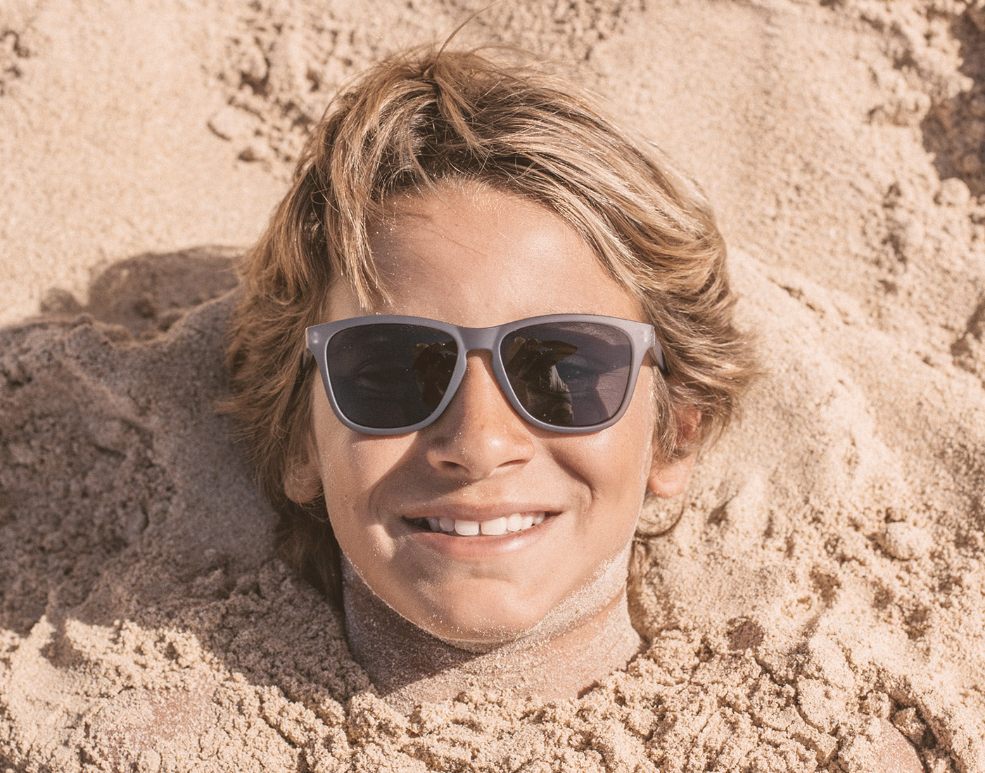 boy buried in the sand wearing sunski mini headland sunglasses