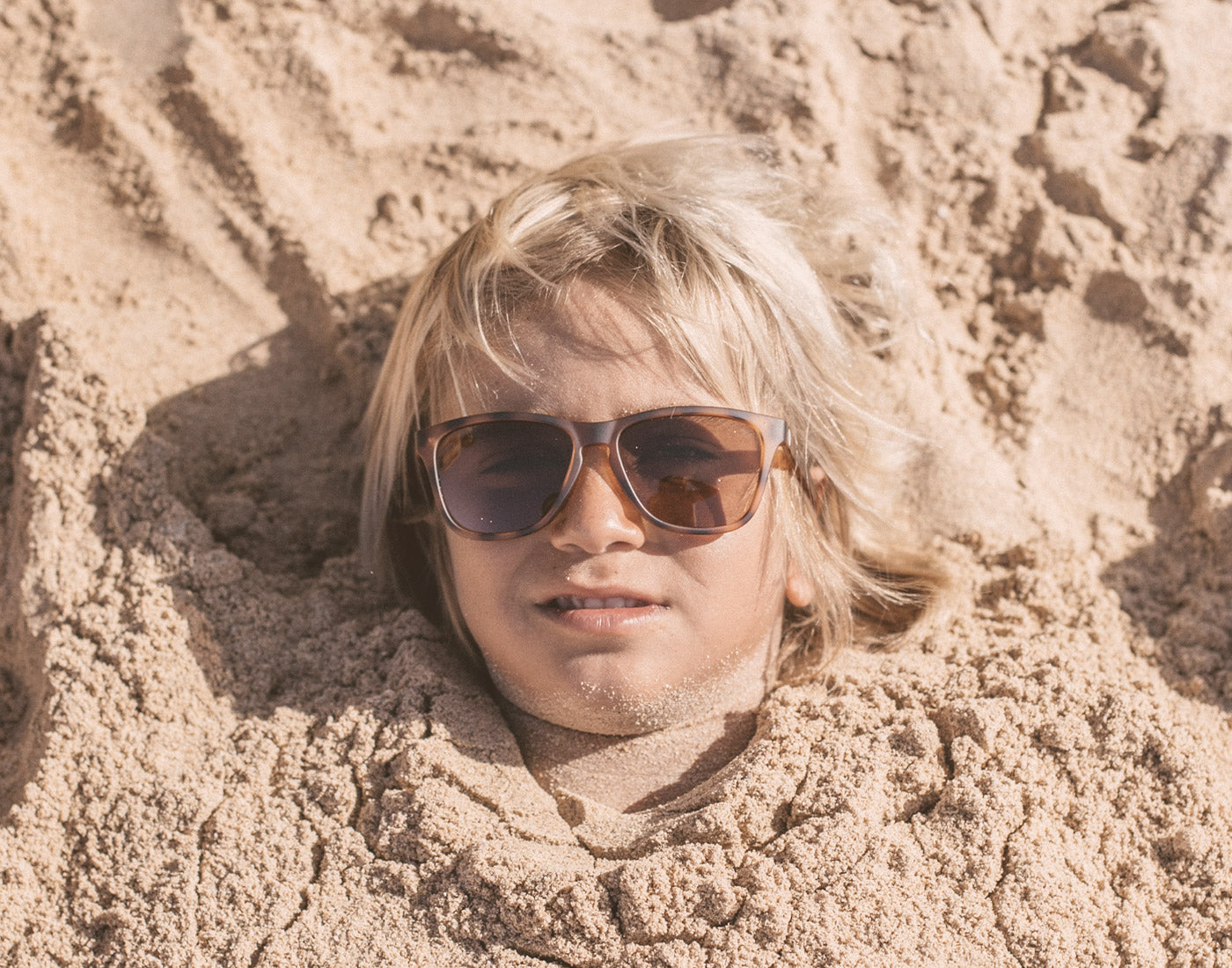 boy buried in the sand wearing sunski mini madrona sunglasses
