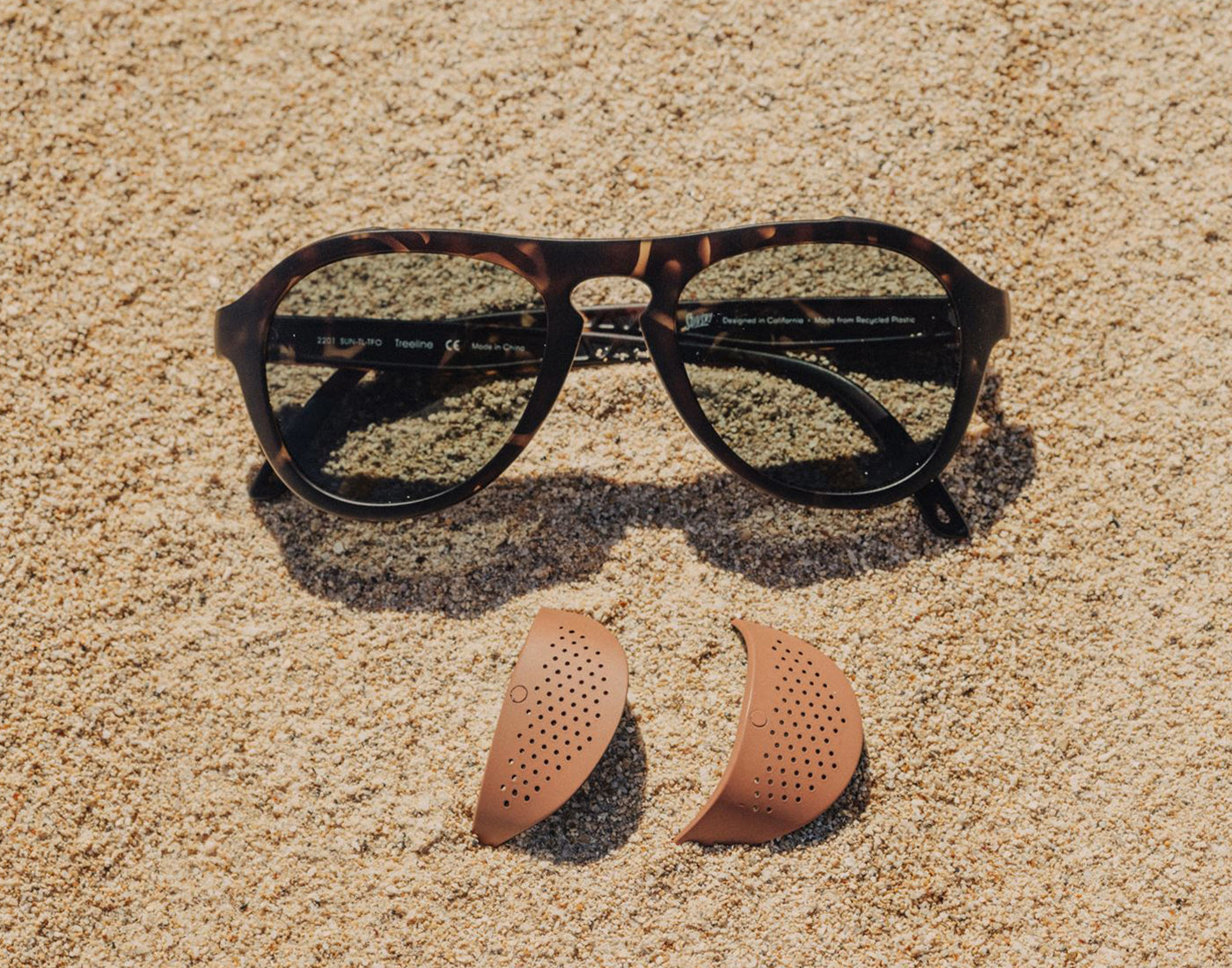 pair of sunski treeline sun shields laying in the sand below sunski treeline sunglasses